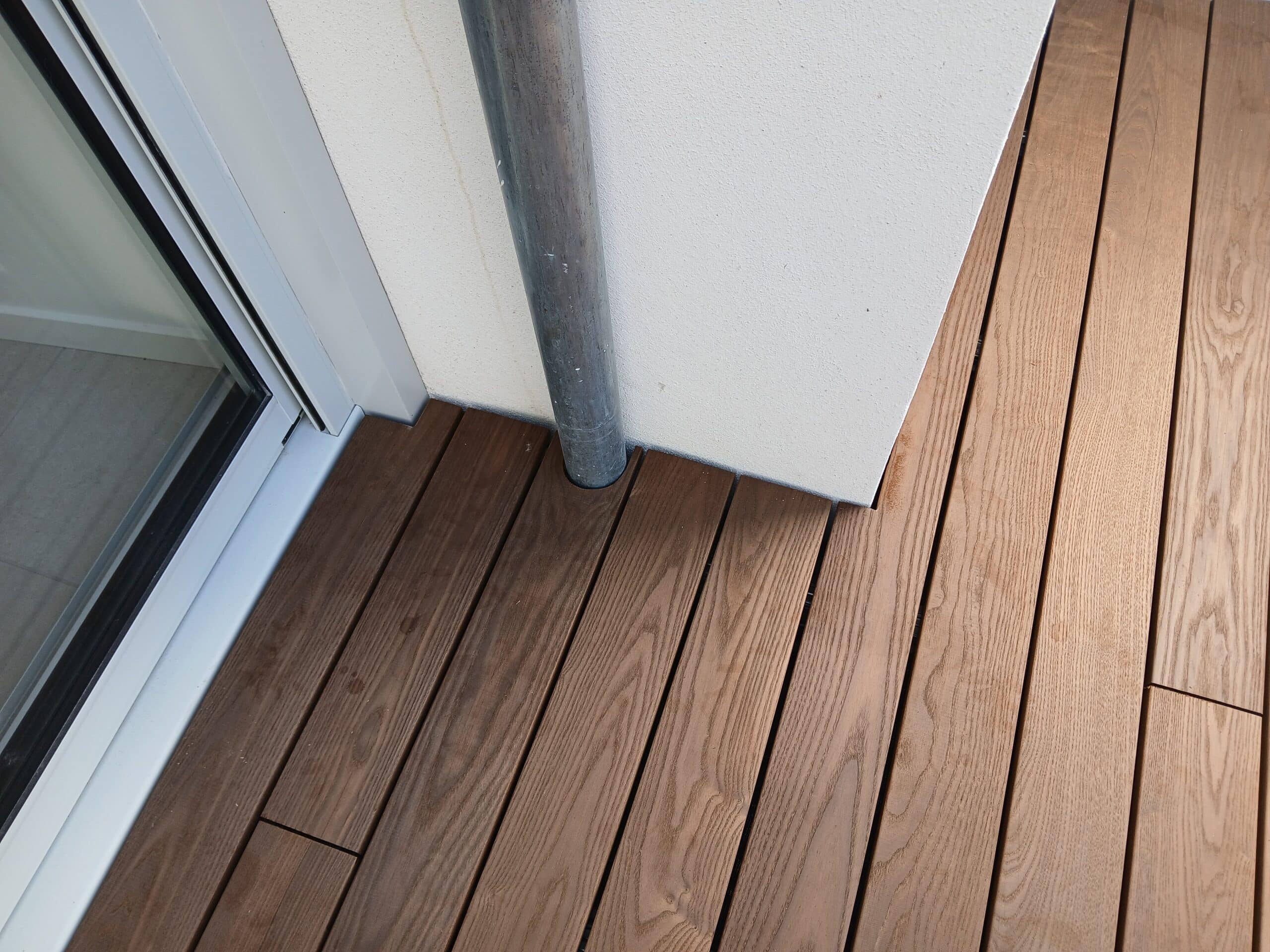 Platelage en bois marron ajusté autour d'un tuyau d'évacuation métallique sur un balcon moderne.
