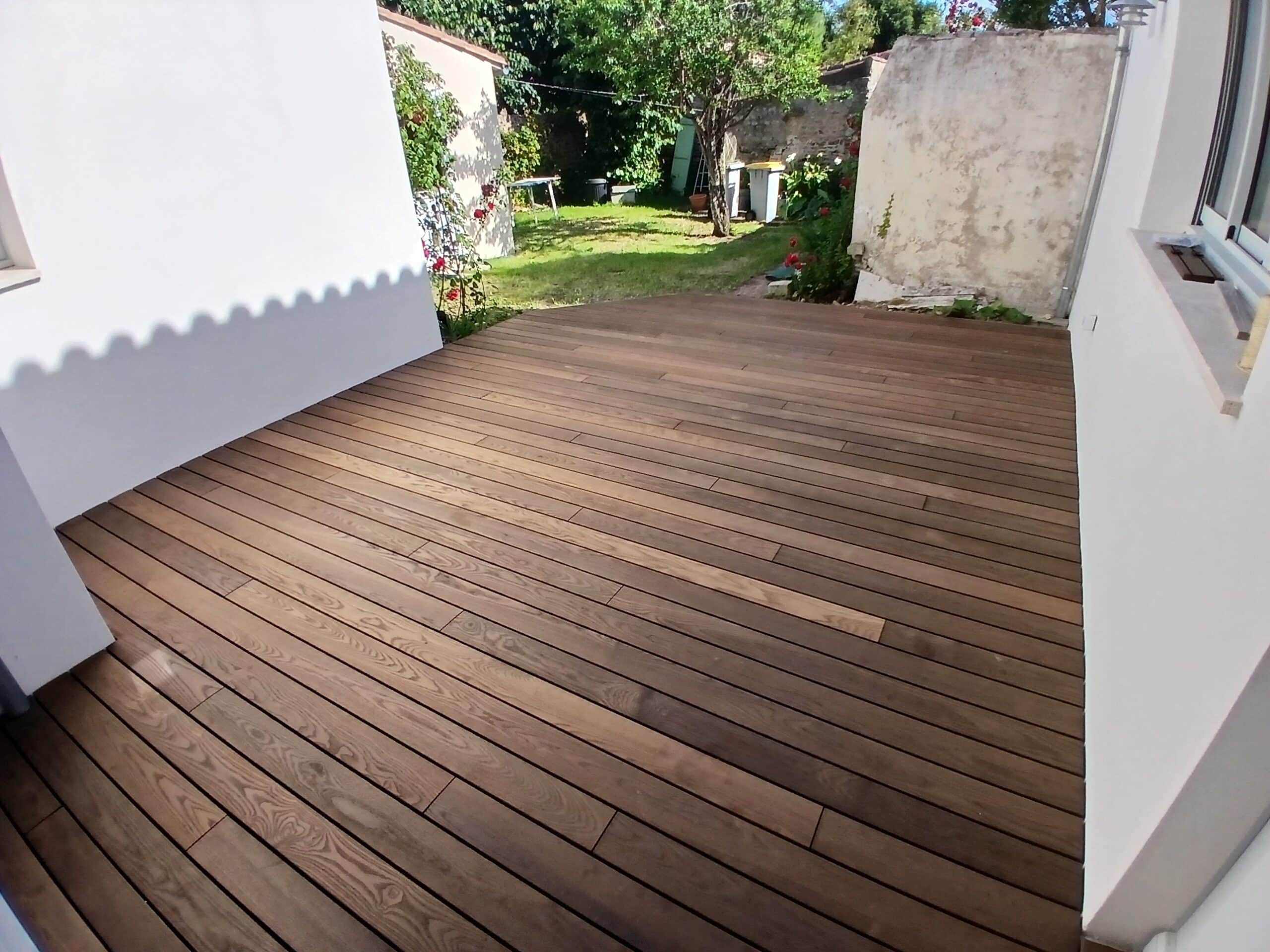 Terrasse en bois brun installée dans une cour entre des murs blancs avec vue sur un jardin verdoyant.