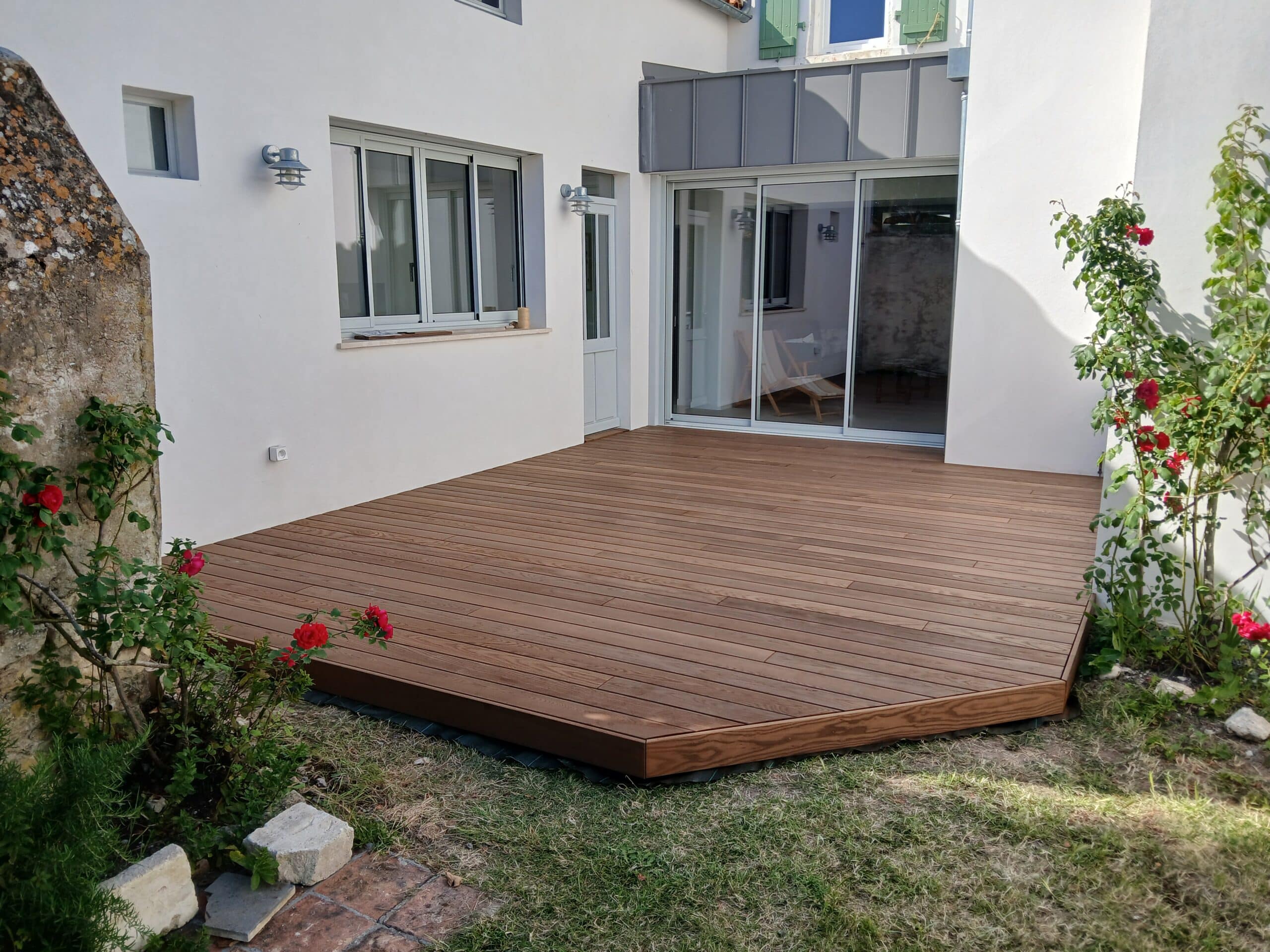 Terrasse en bois exotique au pied d'une maison blanche moderne avec rosiers rouges et large baie vitrée.