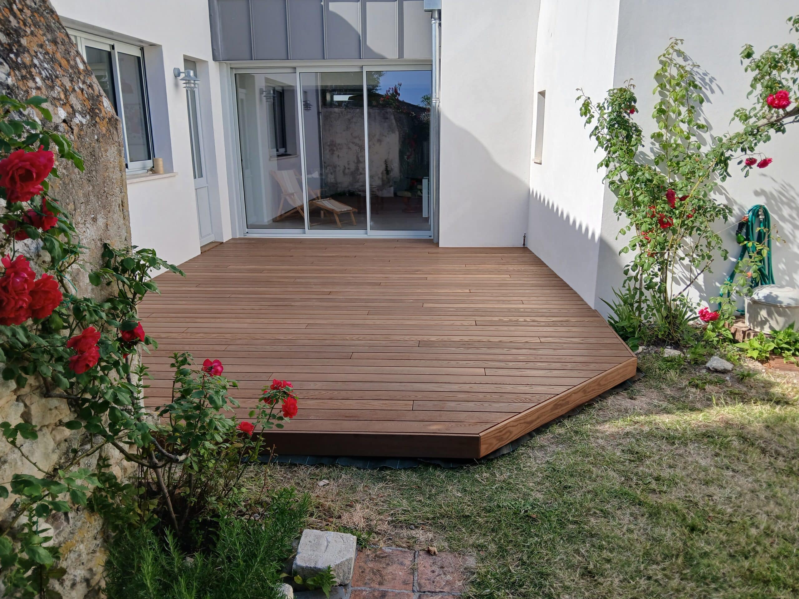 Terrasse en bois clair moderne dans un jardin fleuri de rosiers rouges près d'une baie vitrée coulissante.