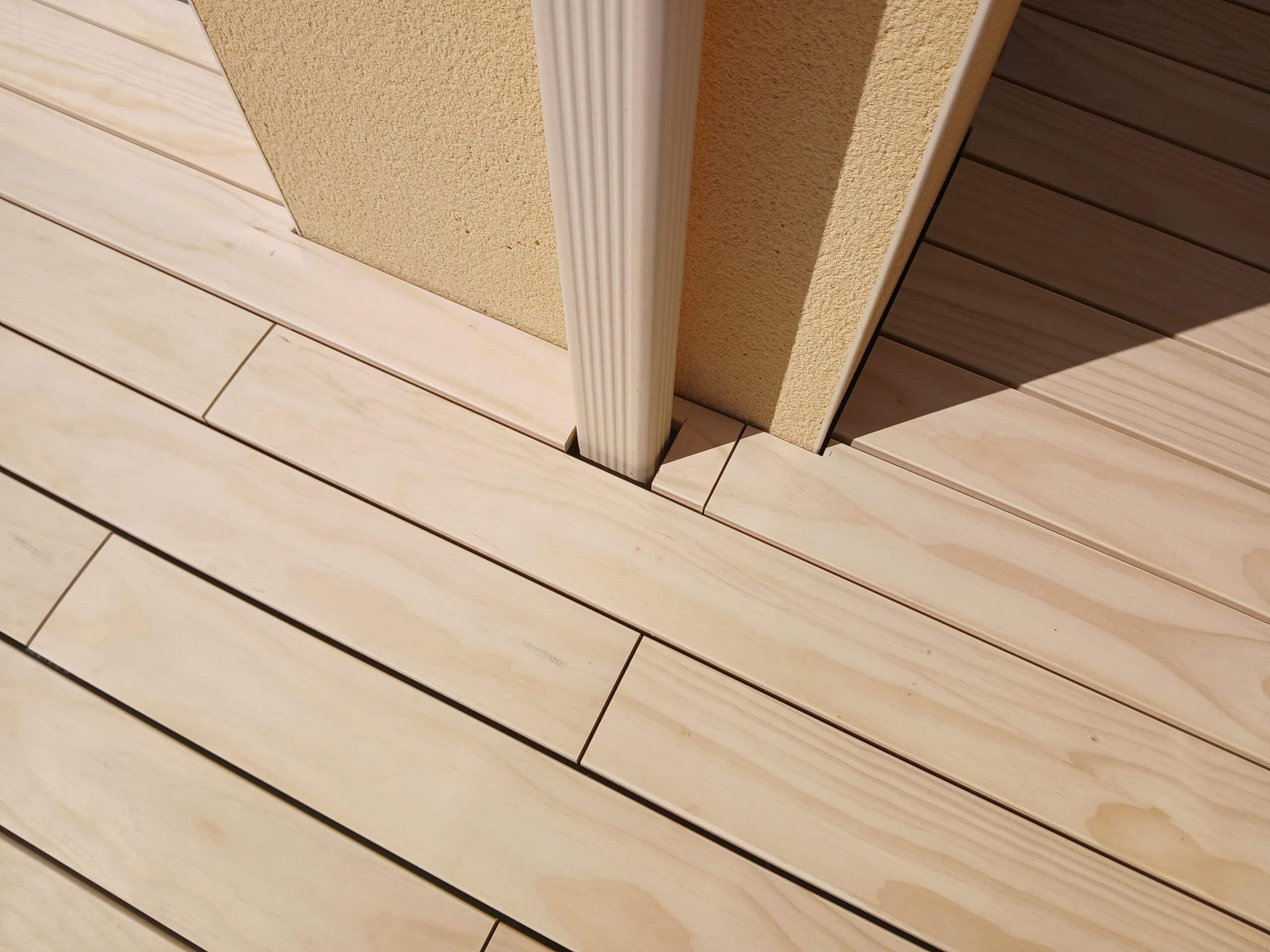 Terrasse en bois clair ajustée avec précision autour d'un mur beige et d'une descente de gouttière blanche.