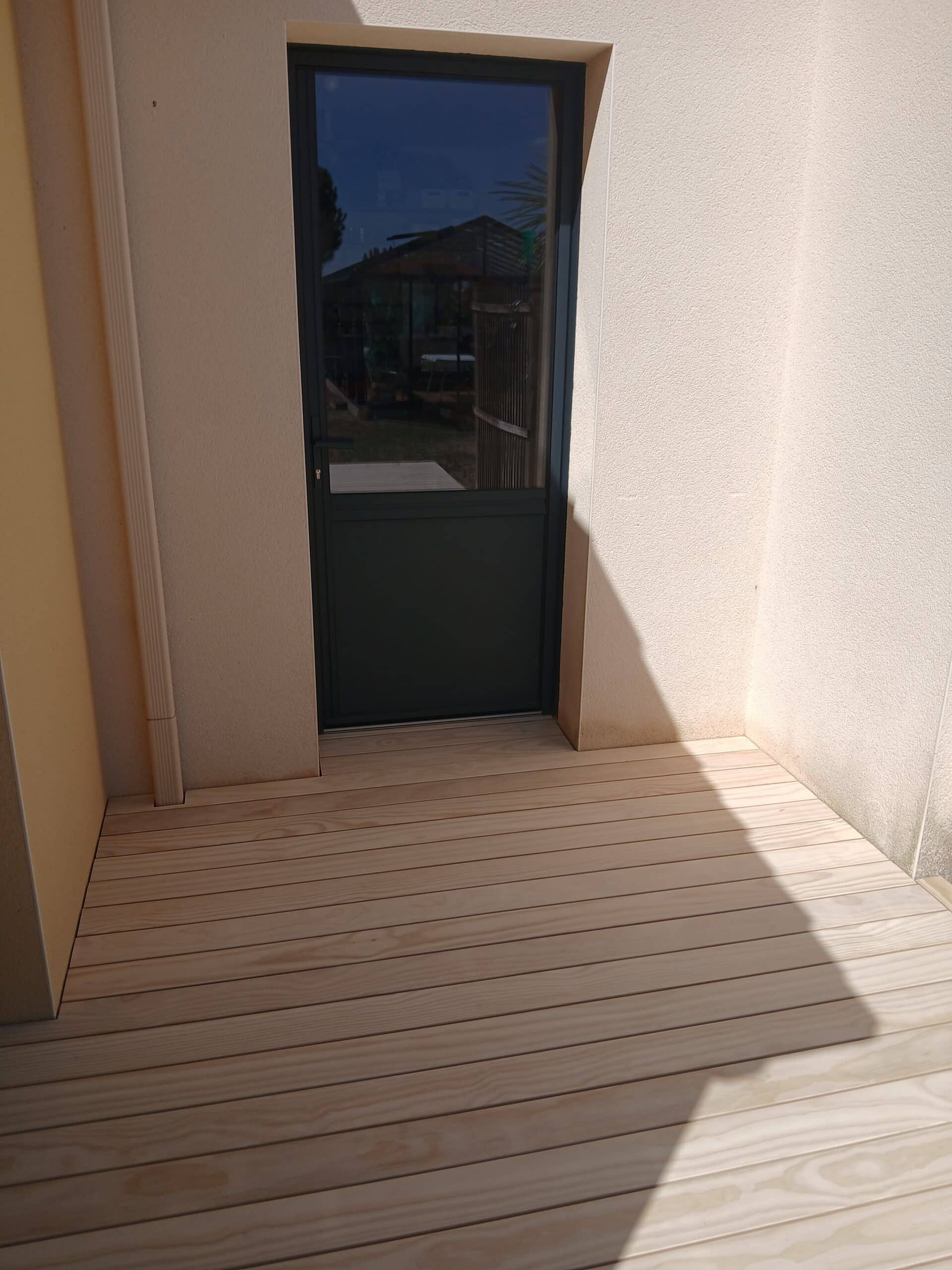 Porte vitrée noire sur terrasse en bois clair et mur beige.