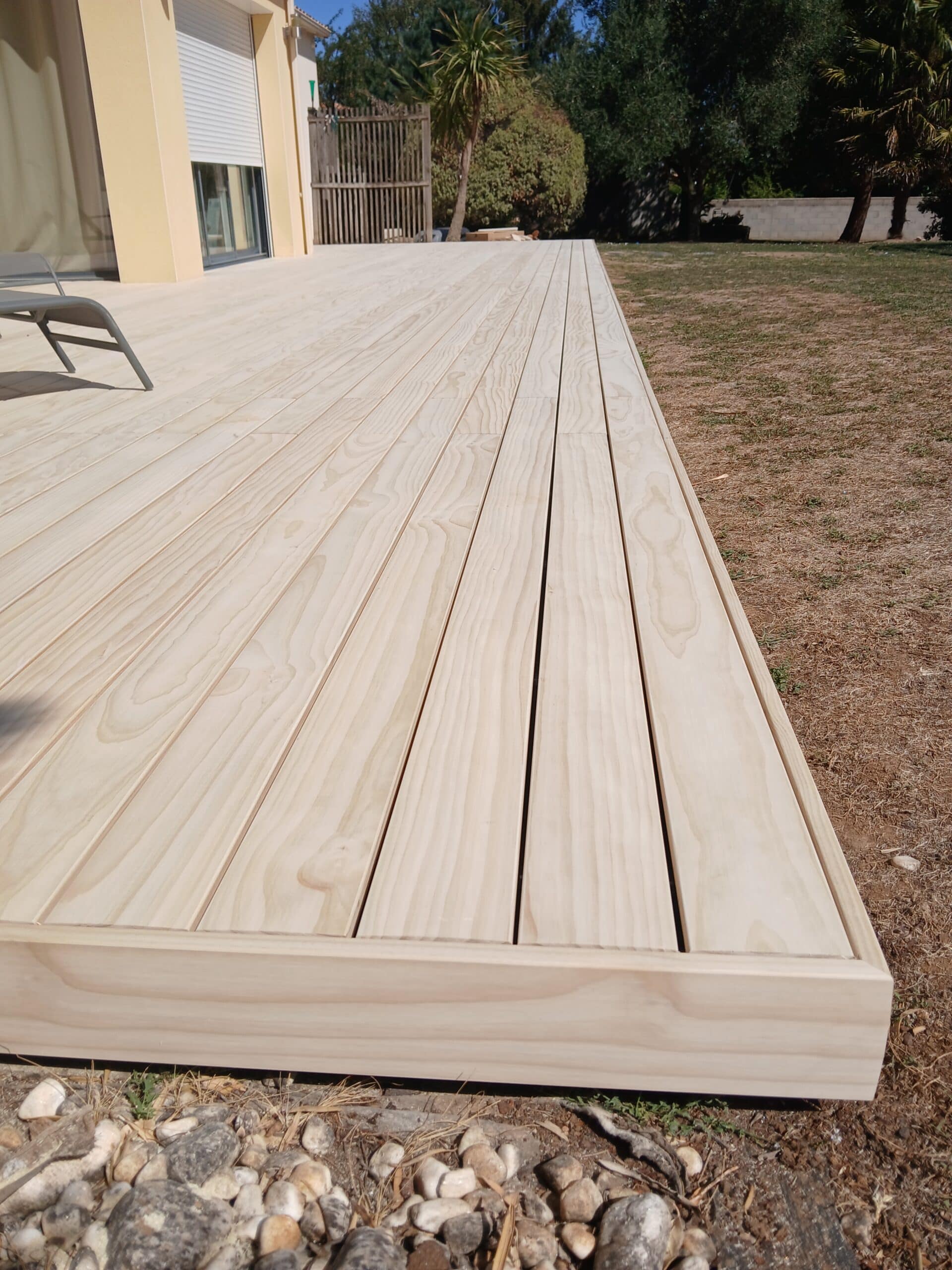 Terrasse en bois clair neuve longeant une maison avec jardin et pelouse sèche.