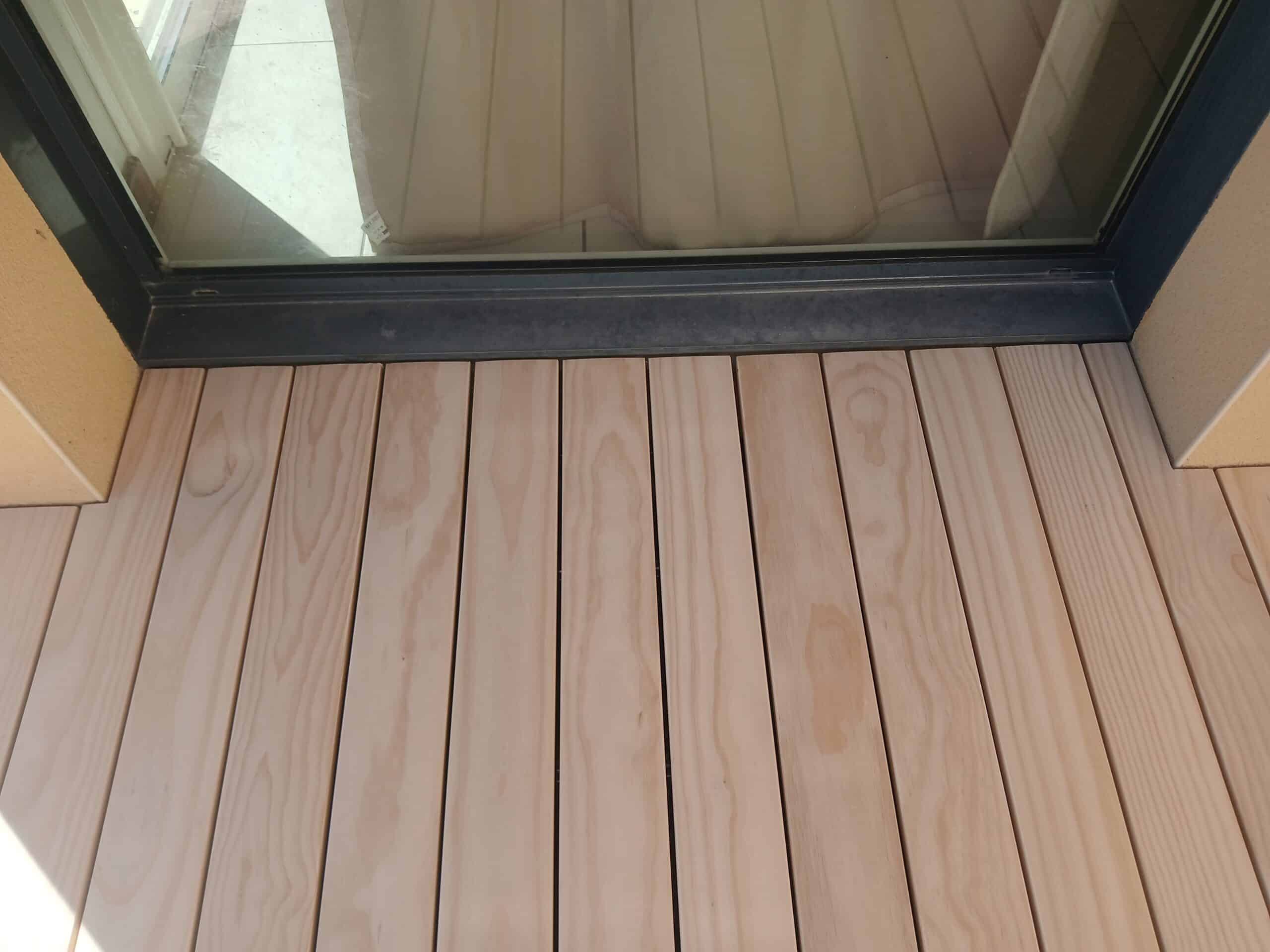 Terrasse en bois clair devant le seuil d'une baie vitrée à cadre noir.
