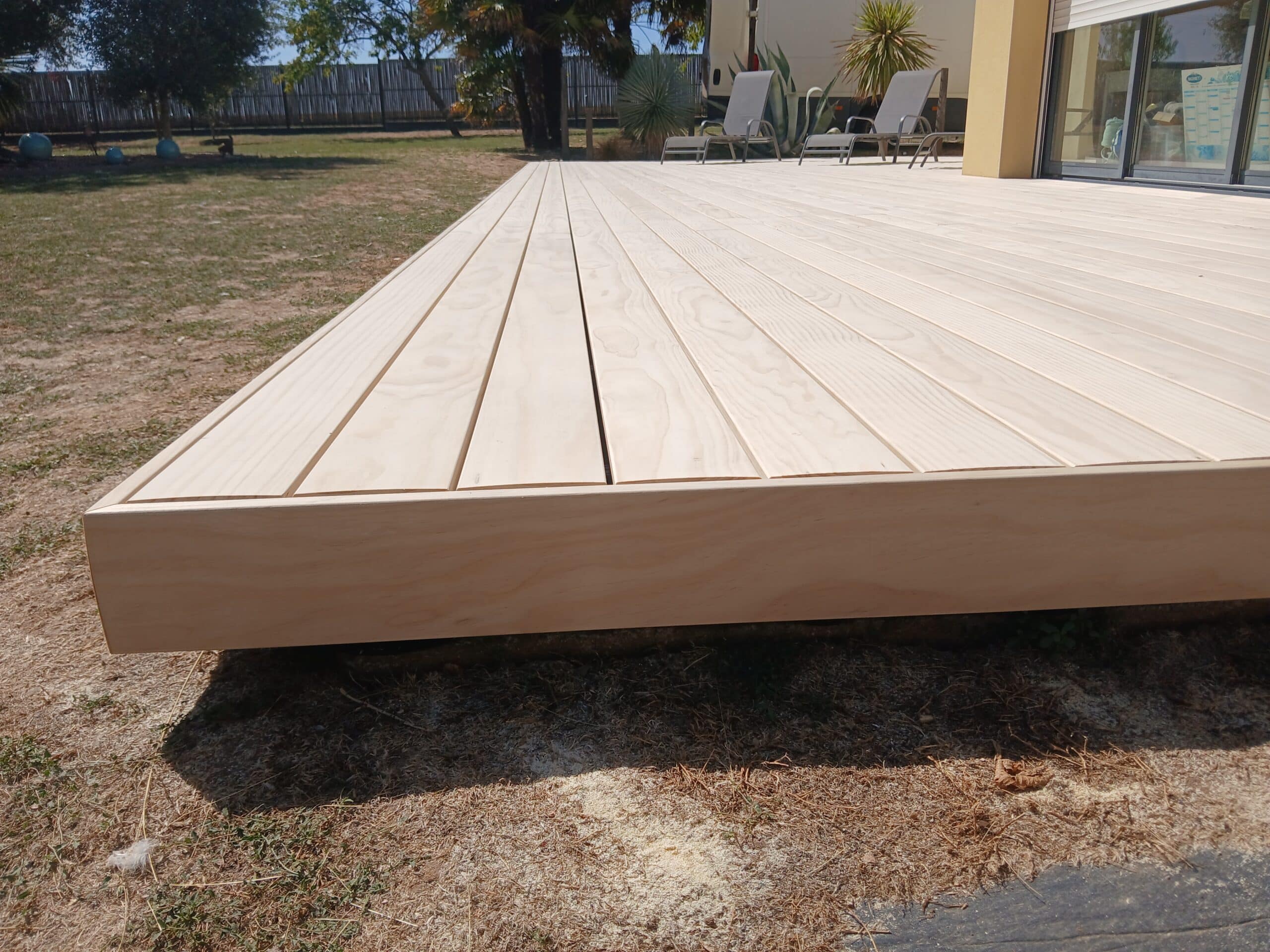 Terrasse en bois clair lisse dans un jardin avec transats en arrière-plan.