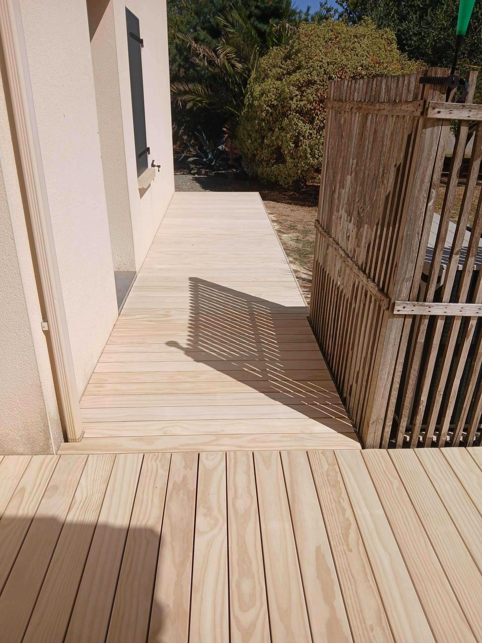 Terrasse en bois clair longeant une façade avec ombres de clôture.