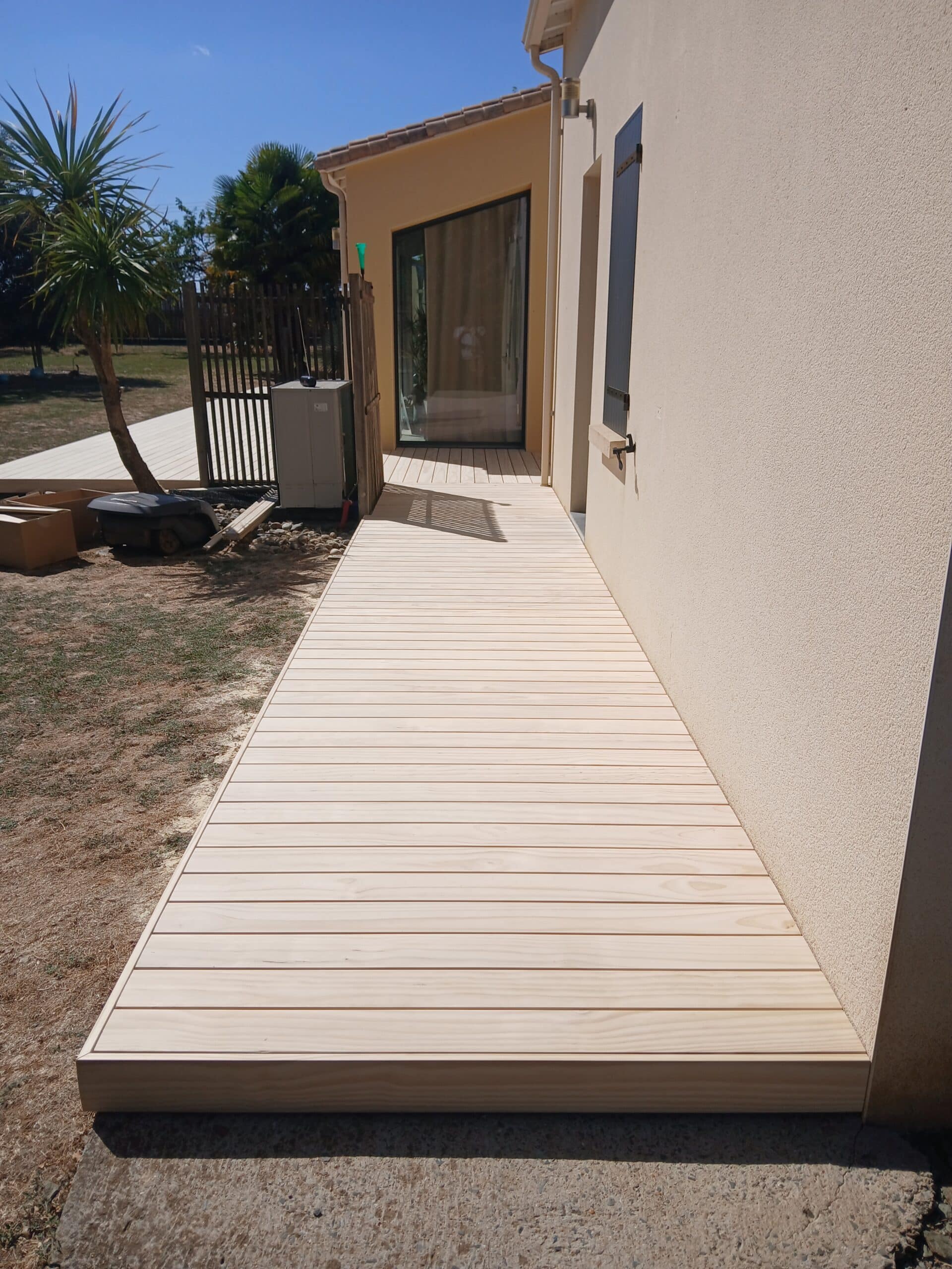 Allée en terrasse bois clair longeant une façade beige, jardin et palmier sous ciel bleu.