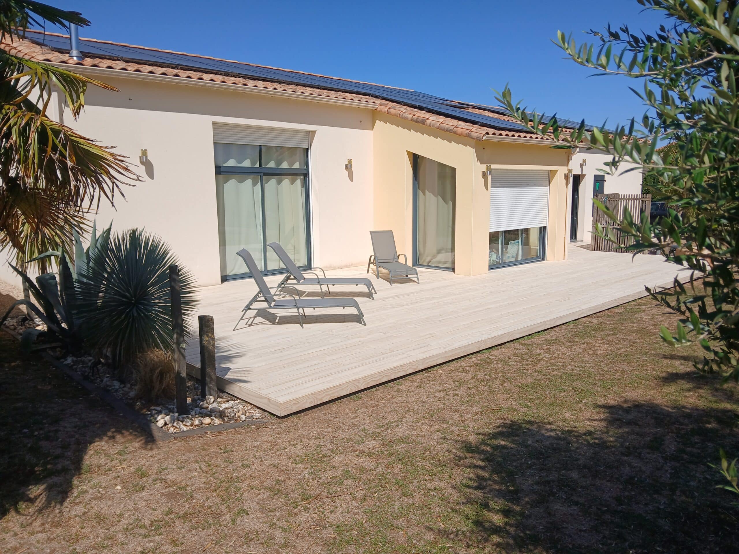 Terrasse en bois clair avec chaises longues devant une maison moderne sous un ciel bleu dégagé.