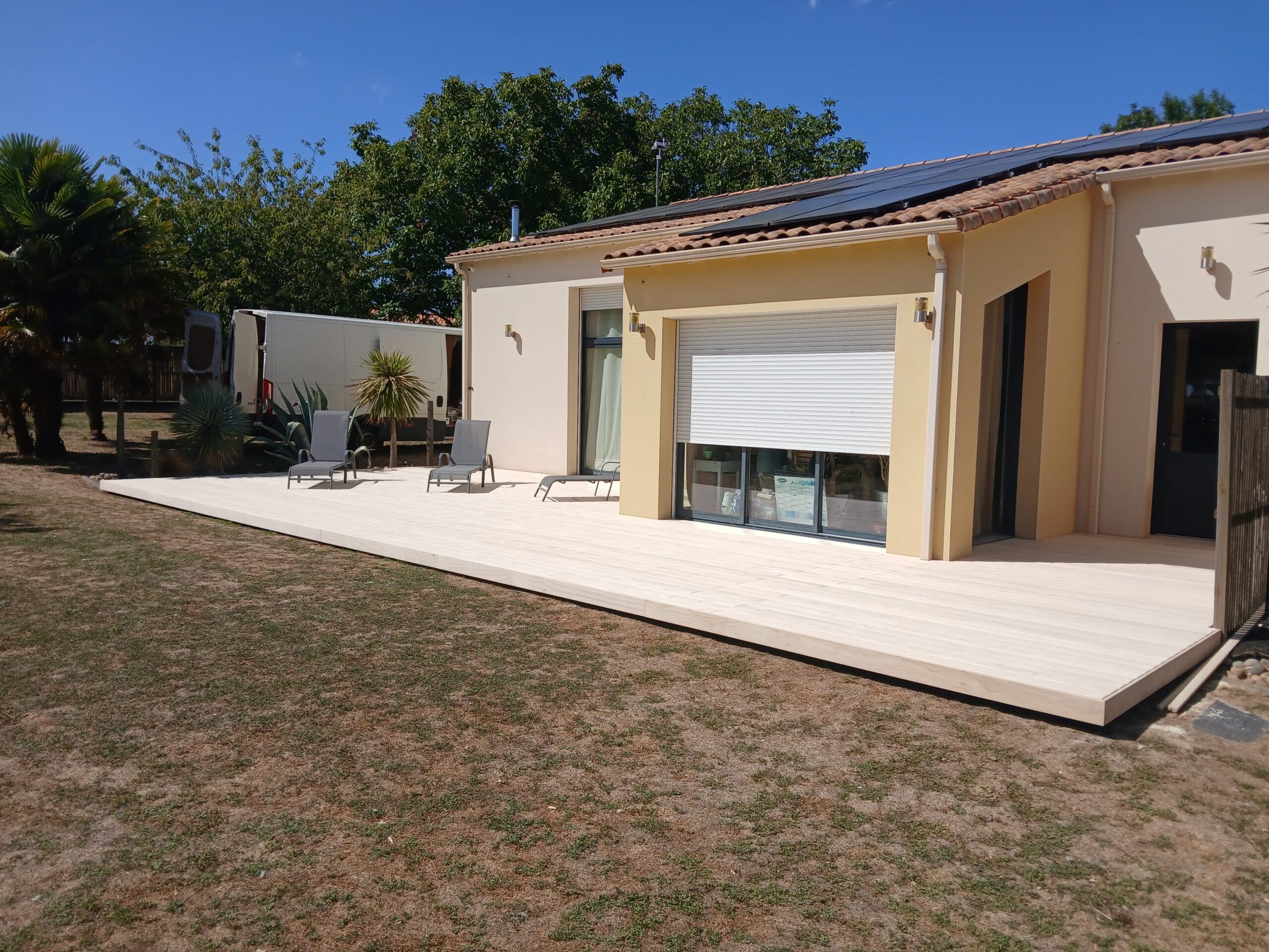 Maison avec terrasse bois, panneaux solaires et transats sous ciel bleu.